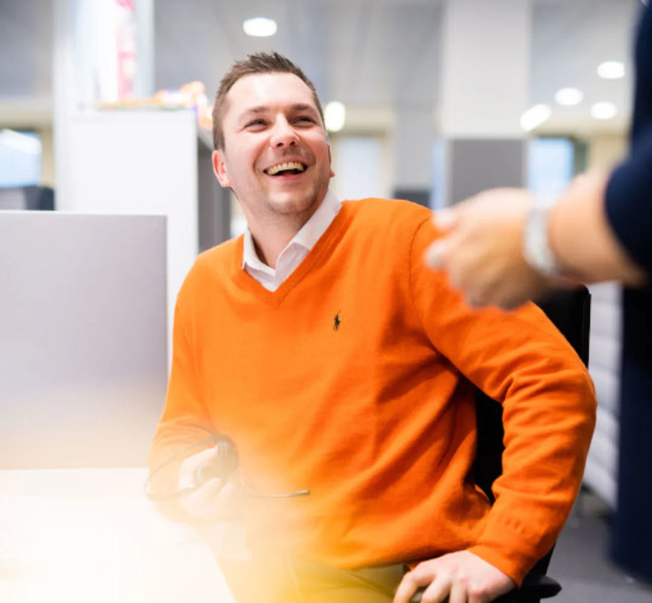 Andreas is laughing with a colleague in the office. He is wearing an orange sweater.