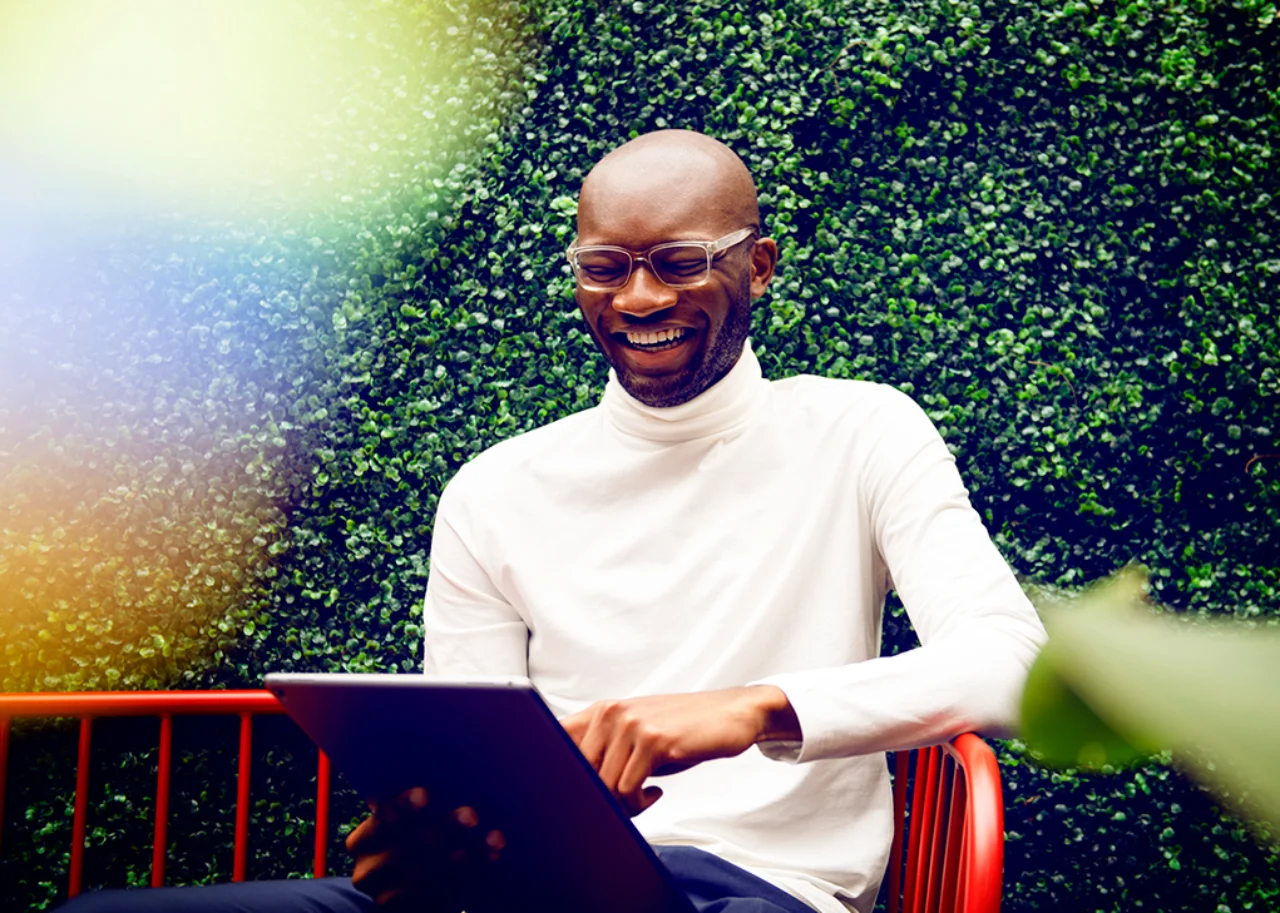 Kofi mit Tablet sitzt lachend auf einer roten Bank vor einer Buchsbaumhecke